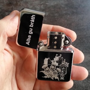 Puede incluir: Un encendedor plateado y negro con el texto "Alba gu bràth" en la tapa. El cuerpo del encendedor presenta un diseño grabado de un castillo, cardos y un gaitero.