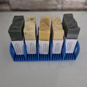 May include: Six bars of soap in a blue soap dish. The soap bars are labeled with the text "The power of nature, cleansing experience, handcrafted for you".