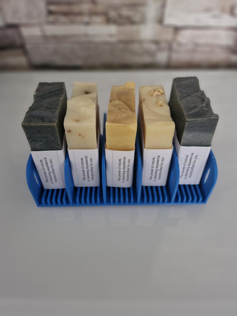 May include: Six bars of soap in a blue soap dish. The soap bars are labeled with the text "The power of nature, cleansing experience, handcrafted for you".