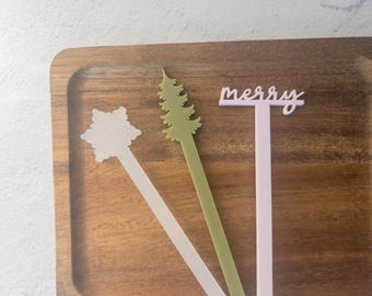 May include: Wooden tray with three decorative drink stirrers. One is a snowflake shape, one is a green Christmas tree, and the third says "merry" in a script font. The stirrers are in white, green, and pink.