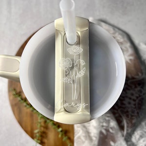 May include: White travel mug with a straw and a floral design on the lid. The lid has a sliding cover to protect the straw. The mug is engraved with the name "Carys".