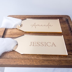 Puede incluir: Dos etiquetas doradas de acrílico con los nombres "Amanda" y "Jessica" grabados. Cada etiqueta está unida a una cinta blanca y descansa sobre una bandeja de madera oscura. Las etiquetas son rectangulares con una parte superior puntiaguda.