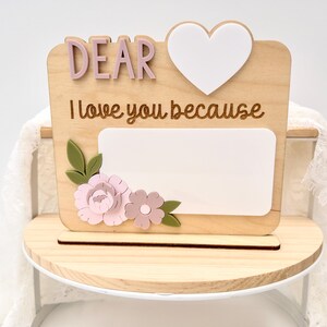 May include: Wooden sign with the words "DEAR" in pink letters and "I love you because" in brown script. A white heart and a blank rectangle are on the sign. Pink and light pink flowers with green leaves decorate the bottom.