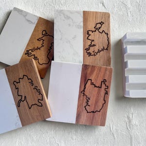 Puede incluir: Un conjunto de posavasos rectangulares con un diseño bicolor. La mitad de cada posavasos es de mármol blanco, y la otra mitad es de madera con un mapa grabado con láser y el nombre del lugar. También hay un jabonera de mármol blanco.