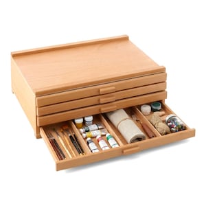 May include: A wooden artist's storage box with multiple drawers, partially open to reveal art supplies. The box is made of light-colored wood and contains paint tubes, brushes, and other art materials. The top of the box has a raised edge.