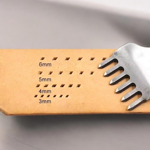 May include: A close-up of a leather craft tool with a metal pronged head. The tool is resting on a piece of tan leather with a grid of holes marked with the measurements 3mm, 4mm, 5mm, and 6mm.