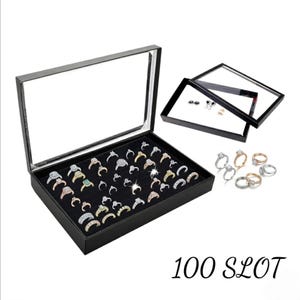 May include: A black display case with a glass lid, filled with various rings. The case is open, revealing a black velvet interior with multiple ring slots. Additional rings are displayed outside the case. The text "100 SLOT" is visible.