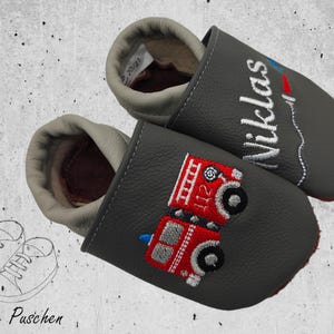 Könnte beinhalten: Ein Paar graue Lederschuhe für Babys mit gestickten Designs. Ein Schuh zeigt ein rotes Feuerwehrauto mit der Nummer 112 und der andere Schuh hat den Namen "Niklas" gestickt.