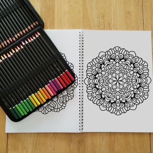 May include: An open colouring book with a black and white mandala design on the right page. A case of coloured pencils is on the left page. The pencils are arranged in rows by colour, with green, yellow, orange, red, pink, and purple pencils visible.