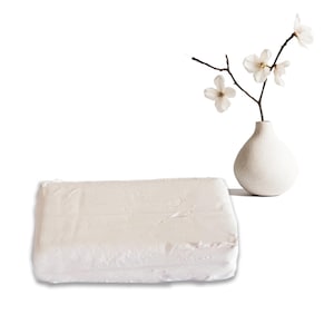Può includere: Una coperta rettangolare bianco sporco con una superficie strutturata. Accanto, un piccolo vaso bianco contiene un ramo con delicati fiori bianchi. Gli oggetti sono posti su uno sfondo bianco, creando un'estetica minimalista.