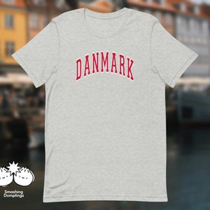 May include: A light heather grey short-sleeve t-shirt featuring the word DANMARK printed in a red collegiate-style font with a white outline across the chest. A small white logo with two dumpling illustrations and the text Smashing Dumplings is visible.