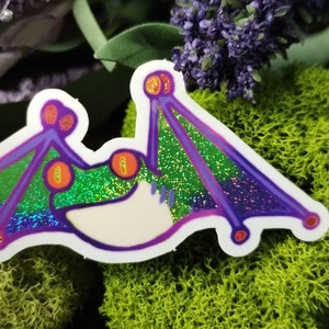 May include: A holographic sticker of a green and purple bat with large, orange eyes. The bat has a white body and is outlined in purple.