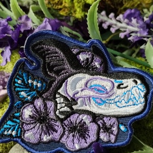 Peut inclure: Patch brodé représentant un crâne avec des détails floraux. Le crâne est blanc avec des accents bleus et violets. Les fleurs sont violettes et bleues avec des accents blancs. Le patch est sur un fond bleu foncé.