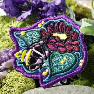 May include: Purple, green, and yellow embroidered patch with a floral design. The patch is made of fabric and has a detailed design with a butterfly and flowers.