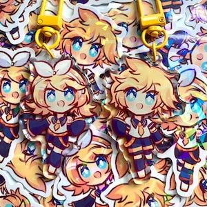 Może przedstawiać: Dwa tęczowe breloki akrylowe z postacią z kreskówki o blond włosach, niebieskich oczach i niebiesko-białym stroju. Breloki są przymocowane do żółtych metalowych klamerek.