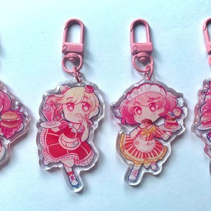 Op de afbeelding: Vier roze en witte acryl sleutelhangers met cartoonfiguren in dienstmeidenoutfits. Elke sleutelhanger heeft een ander personage dat een ander voedsel vasthoudt, waaronder een hamburger, frietjes, een milkshake en een cupcake.