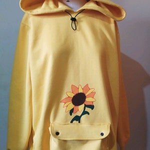 Könnte beinhalten: Ein gelber Hoodie mit einem Sonnenblumenmuster auf der Vorderseite. Der Hoodie hat eine Kängurutasche mit einem Druckknopfverschluss.