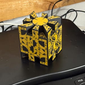 May include: A yellow and black puzzle box with intricate geometric designs. The box has a central circular element and is composed of multiple rectangular panels. The design features sun-like and abstract patterns.
