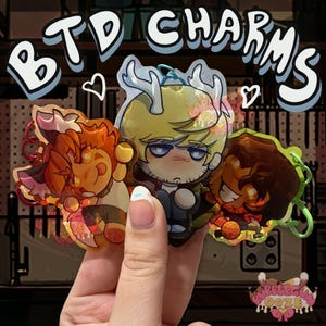 May include: Three acrylic charms labeled BTD CHARMS featuring stylized anime-inspired characters. One charm depicts a blonde character with white antlers, another shows an orange-haired creature, and the third features a dark-haired character.