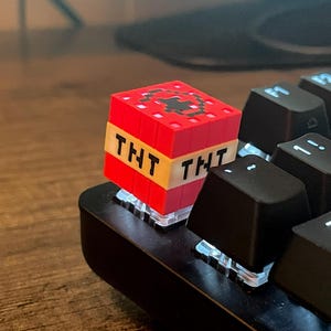 Könnte beinhalten: Ein roter und schwarzer TNT-Block-Keycap mit dem Wort "TNT" auf einem gelben Band. Der Keycap befindet sich auf einer schwarzen Tastatur, mit anderen schwarzen Tasten sichtbar. Das Zubehör hat ein Minecraft-Thema.