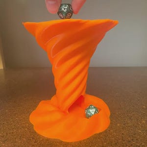 May include: An orange, 3D-printed dice tower in a tornado shape. A hand is placing a green and black 12-sided die on top. Another die rests in the base. The tower has a swirling, textured design.