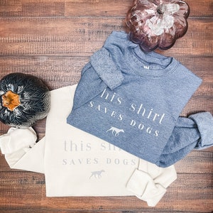 May include: Two sweatshirts, one light beige and one dark blue, with the text "this shirt saves dogs" and "his shirt saves dogs" printed on them. Both sweatshirts feature a small graphic of a dog.