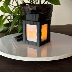 May include: A black Minecraft-style lantern with a glowing orange light inside. The lantern is made of small square blocks and has a clear glass window on each side.