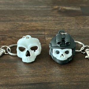 May include: Two keychains with skull designs. One is a white skull, and the other is a black helmet with a skull face. Both have silver key rings and clasps. The keychains are on a dark brown wooden surface.