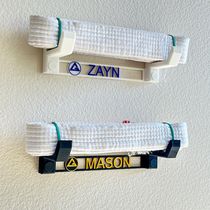 Pode incluir: Dois suportes de parede brancos para cintos com ganchos pretos. O suporte superior tem uma placa de nome branca com um tri&acirc;ngulo azul e o nome "ZAYN". O suporte inferior tem uma placa de nome preta com um tri&acirc;ngulo dourado e o nome "MASON".