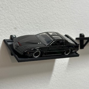 May include: A black die-cast model car of a 1980s sports car, mounted on a black wall shelf. The car has a clear plastic roof and silver wheels.