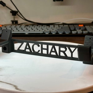 Pode incluir: Suporte de parede preto com o nome "ZACHARY" recortado no centro. O suporte tem dois ganchos de cada lado.