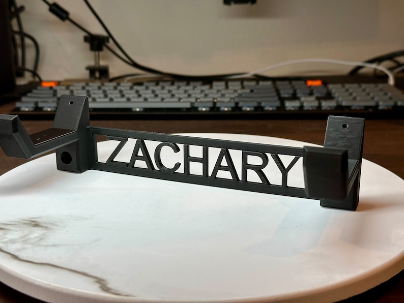 Pode incluir: Suporte de parede preto com o nome "ZACHARY" recortado no centro. O suporte tem dois ganchos de cada lado.