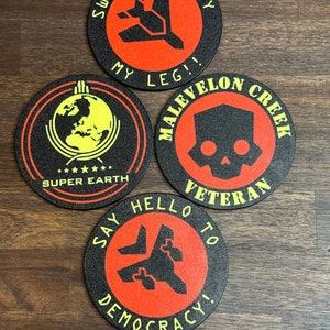May include: Four round black coasters with red and black graphics and text. One coaster says "Sweet Liberty My Leg!!", another says "Super Earth", a third says "Malevelon Creek Veteran", and the last says "Say Hello to Democracy!"