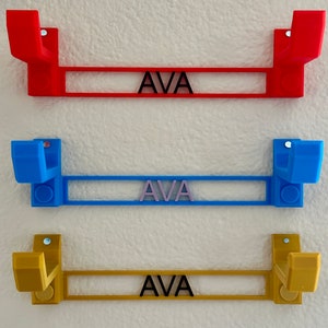 Pode incluir: Tr&ecirc;s ganchos de parede impressos em 3D com o nome "AVA" impresso em cada um. Os ganchos s&atilde;o vermelho, azul e dourado.