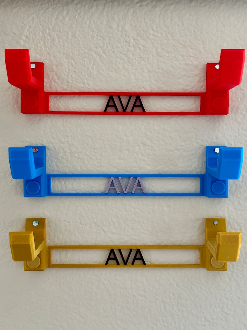 Pode incluir: Tr&ecirc;s ganchos de parede impressos em 3D com o nome "AVA" impresso em cada um. Os ganchos s&atilde;o vermelho, azul e dourado.
