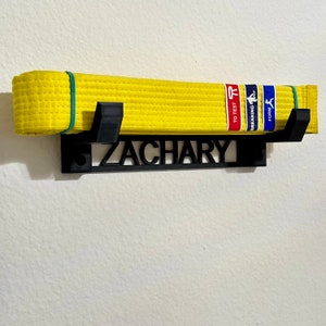 Pode incluir: Suporte de parede preto com o nome "ZACHARY" e dois ganchos para pendurar um cinto de artes marciais amarelo com uma etiqueta azul e vermelha que diz "BREAKING TO TEST FORM".