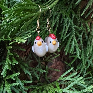 May include: A pair of handmade earrings featuring small, white, bird-shaped charms with red crests and yellow beaks. The earrings hang from gold-colored hooks. The background is a backdrop of green foliage.