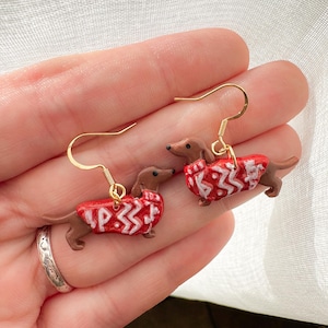 May include: A pair of dachshund earrings. Each earring features a brown dachshund wearing a red sweater with a white pattern. The earrings have gold-colored hooks.