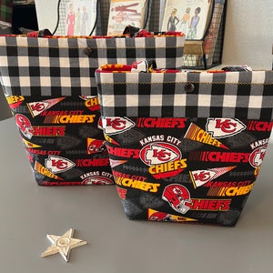 Könnte beinhalten: Zwei schwarze und weiße karierten Tragetaschen mit einem Kansas City Chiefs Muster. Die Taschen haben rote, gelbe und weiße Akzente. Das Muster beinhaltet das Teamlogo, die Worte "Kansas City Chiefs" und Fußbälle.