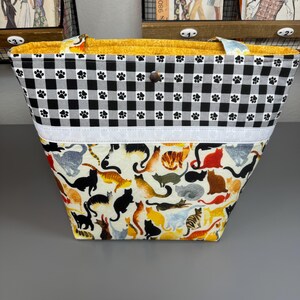 Op de afbeelding: Een zwart-wit geblokte tote bag met poot afdrukken. De tas heeft een witte streep en een onderpaneel met een kleurrijke kattenprint.