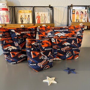 May include: Three different sized tote bags featuring a Denver Broncos theme. The bags are made of fabric and have a blue and orange colour scheme with the Denver Broncos logo and text. The bags are sitting on a grey surface.
