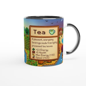 May include: A white ceramic mug with a silver rim and handle. The mug features a colourful design from a video game, including the word "Tea" and a description of its effects. The background shows a landscape with trees, water, and a house.