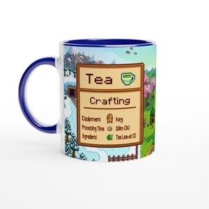 May include: A white ceramic mug with a blue interior and handle. The mug features a colourful design with the text "Tea Crafting" and a pixelated tea cup icon. The design includes a snowy mountain scene and a green landscape.