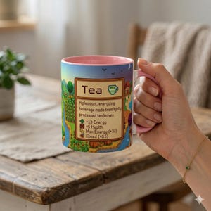 May include: A ceramic mug with a pink interior and handle, displaying a colorful design with the word "Tea" and its effects. The mug is held against a blurred background.