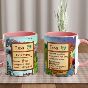 May include: Two white ceramic mugs with pink handles and interiors. Each mug features a colourful design with the word "Tea" and a description. One mug says "Crafting" and the other says "A pleasant, energising beverage made from lightly processed tea leaves."