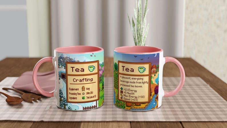 May include: Two white ceramic mugs with pink handles and interiors. Each mug features a colourful design with the word "Tea" and a description. One mug says "Crafting" and the other says "A pleasant, energising beverage made from lightly processed tea leaves."