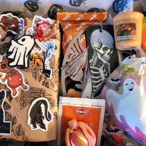 May include: A Halloween-themed gift box filled with various items. Includes stickers, candy, a candle, and a ghost-shaped bag. The stickers feature spooky characters and phrases. The box is decorated with Halloween-themed wrapping paper.