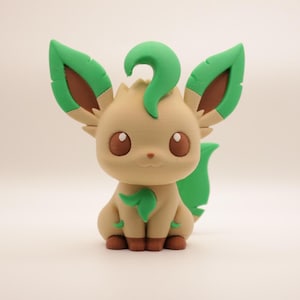 May include: A 3D-printed figure of a Leafeon Pokémon. The figure is primarily tan with green accents on the ears, tail, and leaf-like features. It has large brown eyes and a small, smiling mouth. The figure is sitting.