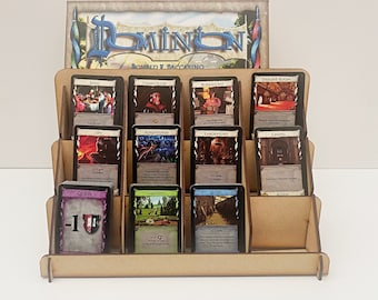 Puede incluir: Un organizador de juegos de cartas de madera con un acabado marrón. El organizador tiene tres estantes y contiene varias cartas de juego de cartas Dominion. Las cartas están dispuestas en filas e incluyen nombres como "Festín", "Canciller", "Burócrata", "Sala del Trono", "Espía", "Aventurero", "Laboratorio", "Capilla", "Maldición", "Jardines", "Foso", y "Acción".