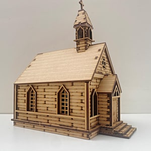 Może przedstawiać: Drewniany model kościoła z wieżą i krzyżem na szczycie. Kościół wykonany jest z jasnobrązowego drewna i ma brązowy dach. Kościół ma okna i drzwi. Model stoi na białym tle.